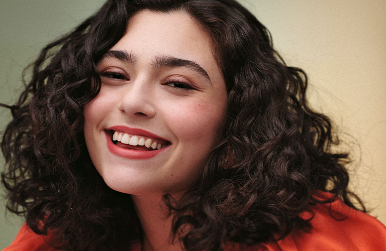 Foto de una mujer con cabello ondulado, largo y con mechas en tonos castaños y rubios. El fondo es amarillo claro, y lleva una blusa beige con textura delicada.